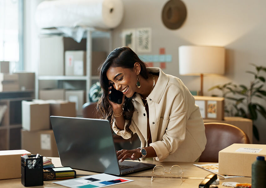 Woman in a beige jacket using a laptop and talking on the phone, surrounded by boxes.
