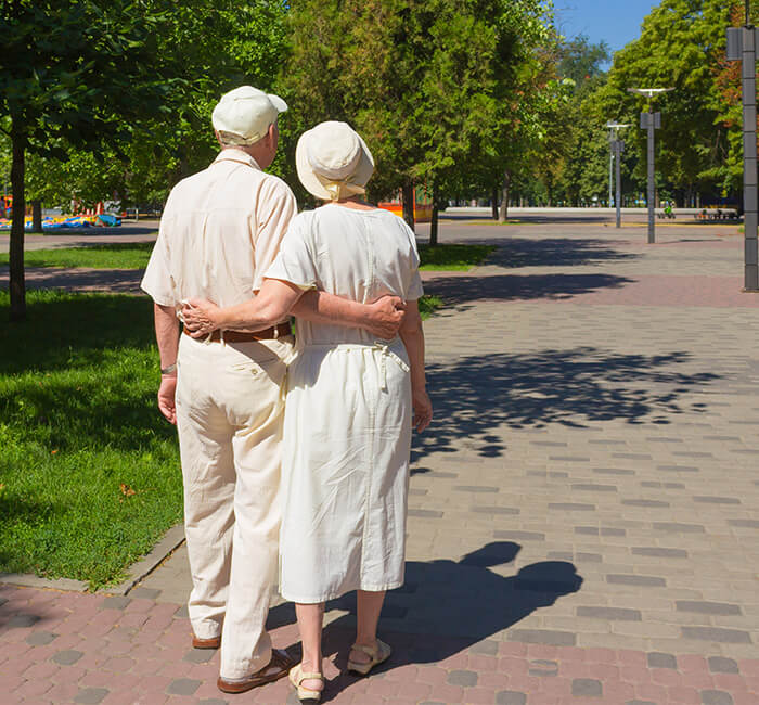 Elderly couple walking arm in arm through a sunny park with green trees.