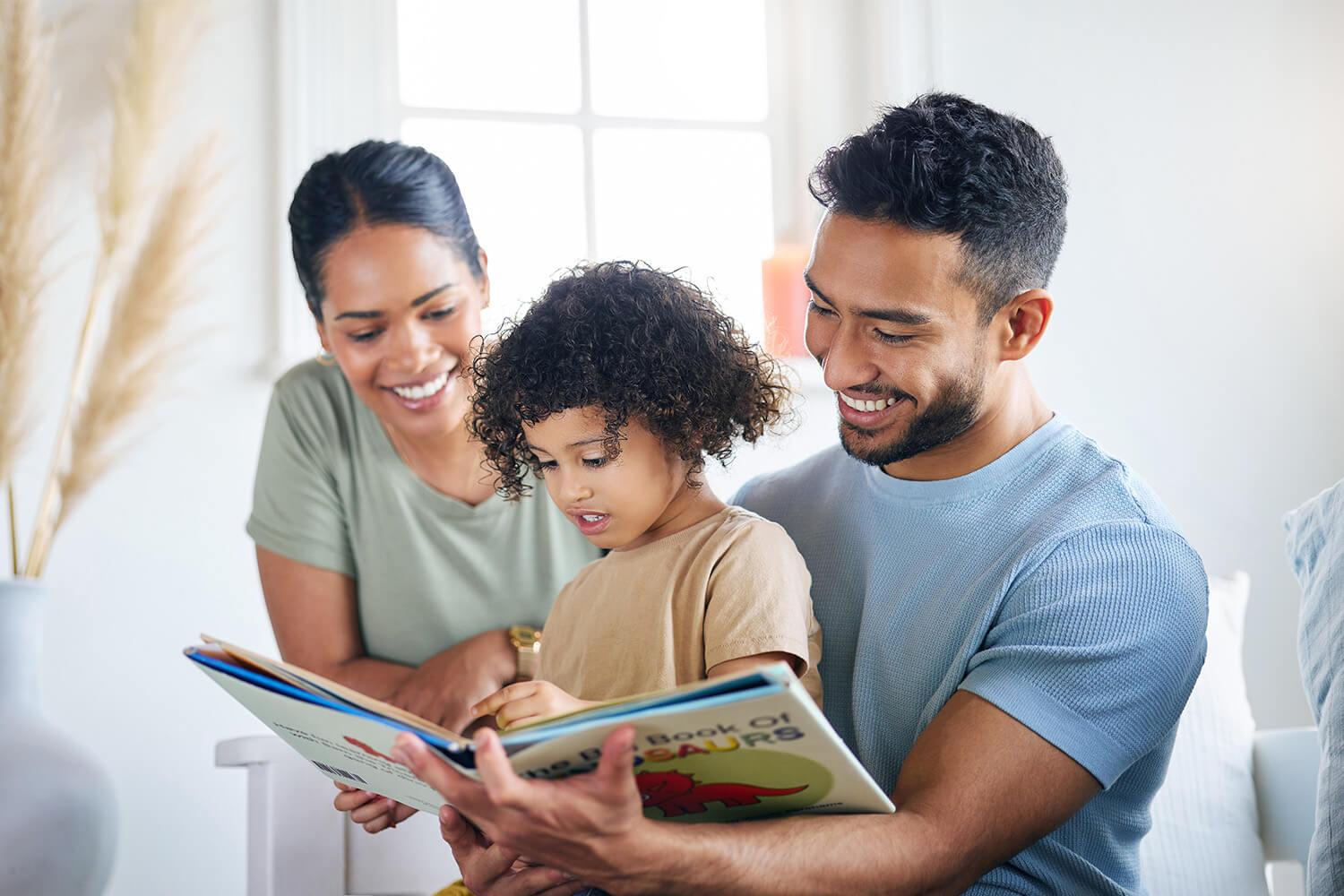Family reading a dinosaur book together near a window.