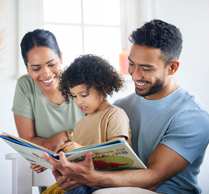 Family reading a dinosaur book together on a bright day indoors.