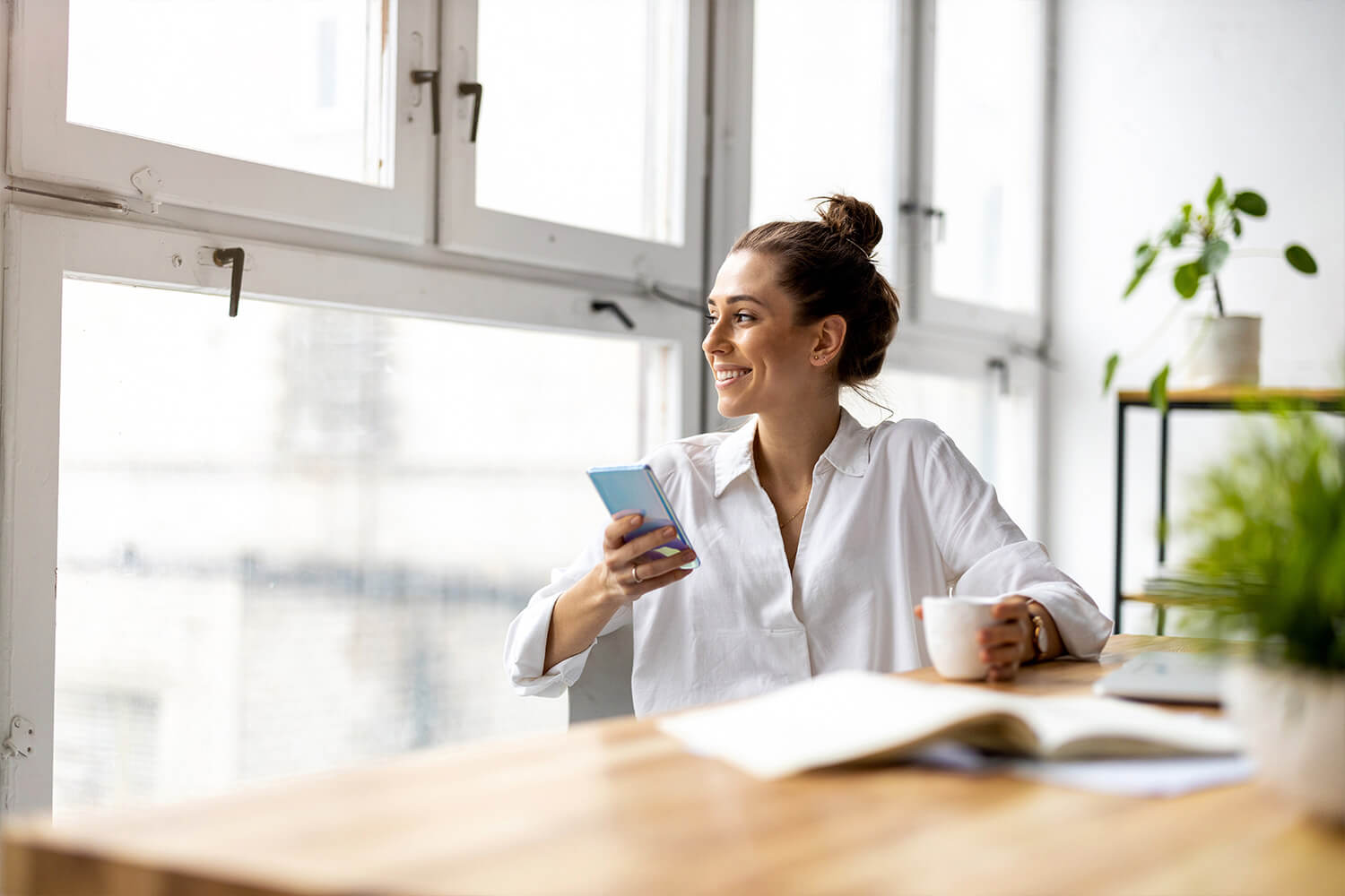 Person in white shirt holding phone by a window, smiling, with a coffee cup on desk.