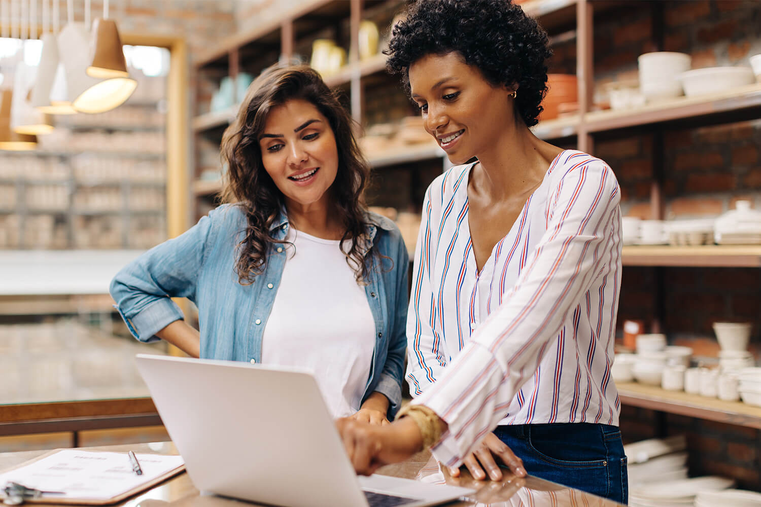 Two women smiling and collaborating at a laptop in a cozy room with shelves.